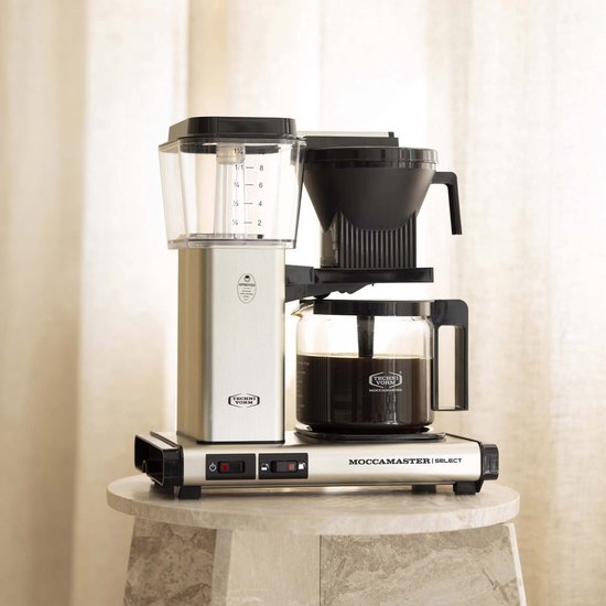 Zilverkleurige Moccamaster Select koffiezetapparaat op marmeren plateau met glazen kan en koffie.