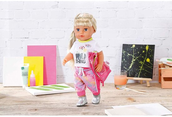 BABY born pop met blond haar, draagt wit shirt met 'WILD THING' opdruk, roze met gele broek en zilveren schoenen. Ze draagt een roze tas over haar schouder.