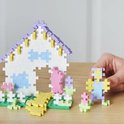 Huisje met paarse dakrand, blauwe ramen, bloemen en een figuurtje met geel, paars en blauw, vastgehouden door een hand.