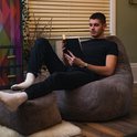 Man leest een boek in een lila beanbag stoel, met zijn voeten op een bijpassende poef. Zittend op een houten vloer.