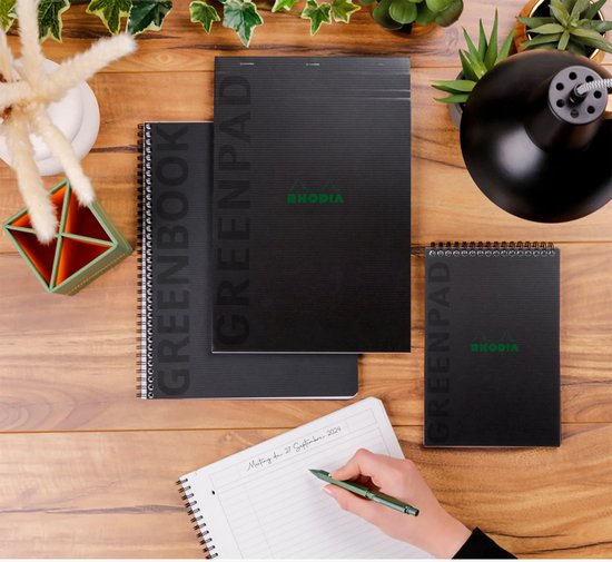 Drie zwarte Rhodia Greenbook notitieboeken in verschillende formaten, op een houten bureau met een schrijfhand.