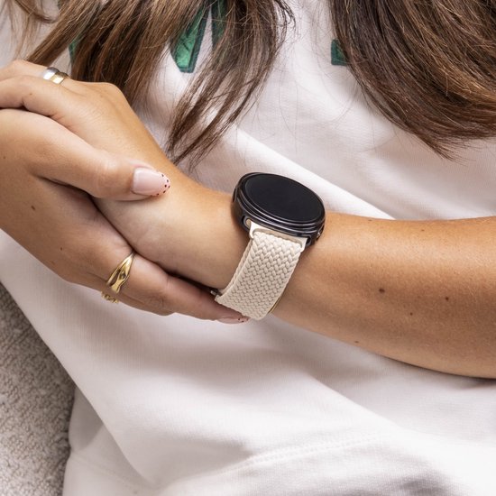 Model met een crème gevlochten smartwatchband om haar pols met een zwarte smartwatch. Een gouden ring en ringen met een parelmoer afwerking.