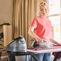 Vrouw met rood shirt en spijkerbroek, strijkt met een zwart Braun CareStyle 3 strijkijzer over een rood textiel. Zwarte stoomgenerator ernaast.