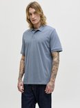 Man met lang blond haar, draagt een blauwgrijze polo en spijkerbroek. Volledige body shot.