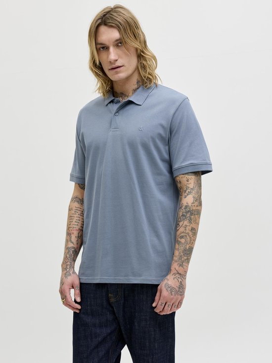 Man met lang blond haar, draagt een blauwgrijze polo en spijkerbroek. Volledige body shot.