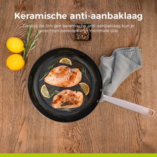 Een koekenpan met gebakken kip, citroen en rozemarijn op een houten tafelblad. Keramische anti-aanbaklaag.