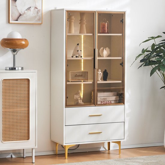 Witte vitrinekast met glazen deuren, gouden handgrepen en gouden poten. Gedecoreerd met beeldjes, een radio en boeken.