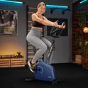 Vrouw op een marineblauwe hometrainer, met fitnessapparatuur op de achtergrond.