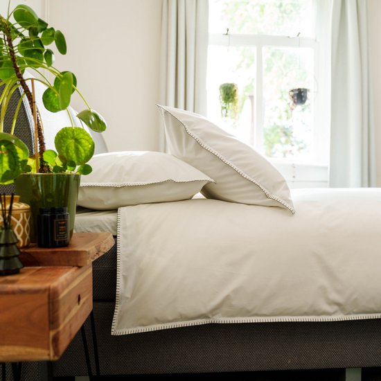 Een wit dekbedovertrek en twee kussenslopen met zigzagstiksels op een bed, naast een nachtkastje met plant.