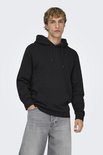 Man met blond haar staat met handen in de zakken van een zwarte hoodie en grijze jeans.
