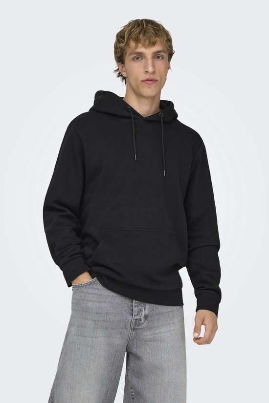 Man met blond haar staat met handen in de zakken van een zwarte hoodie en grijze jeans.