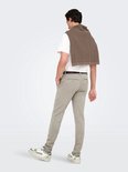 Man in een beige chino, wit T-shirt en bruine trui over de schouders, met witte sneakers.
