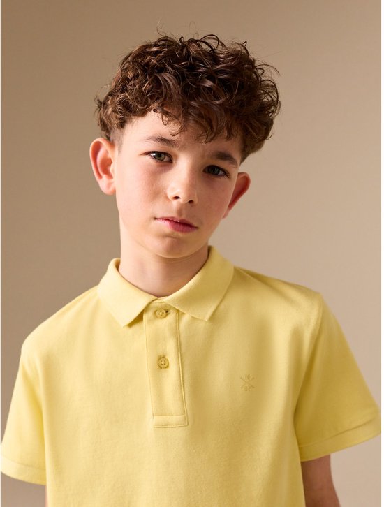 Jongen met krullend haar draagt een lichtgeel poloshirt met korte mouwen en een geborduurd logo.