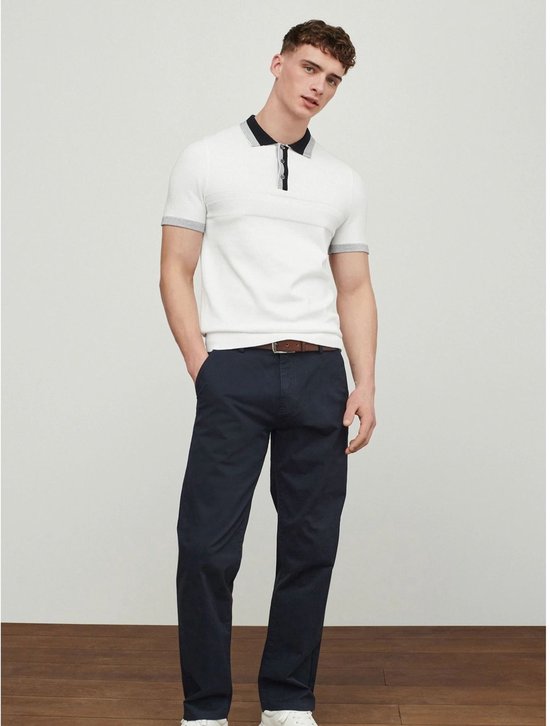 Model in blauwe chino broek met bruine riem en wit poloshirt met grijze details.