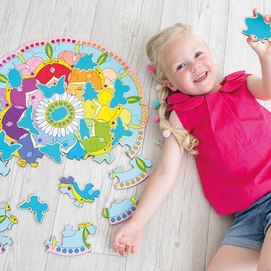 Une fille souriante aux couettes blondes avec des pompons est allongée sur le sol à côté d'un puzzle rond, avec des pièces détachées de chevaux et de bots. Le puzzle est un mandala avec des fleurs.