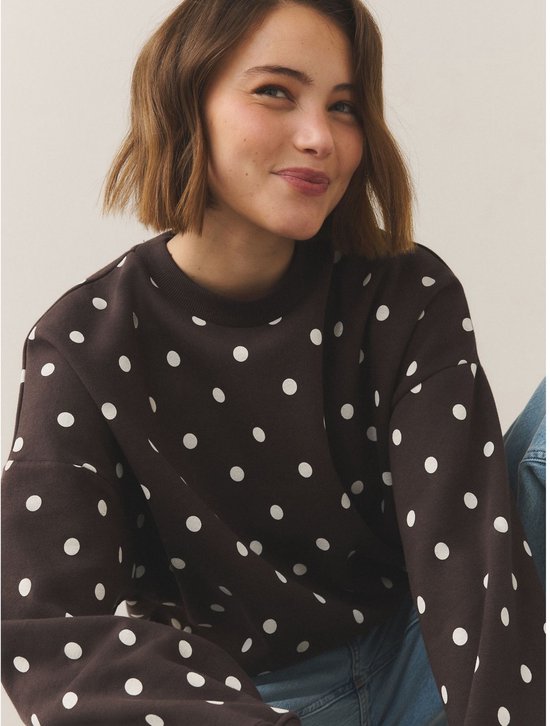 Model met korte bruine bob glimlacht naar links in een donkerbruine sweater met witte polkadotprint.