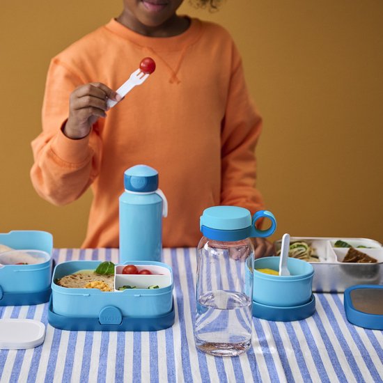 Kind met oranje trui prikt tomaatje met vork. Twee blauwe Mepal lunchboxen en fles staan op tafel.