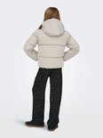 Meisje met blonde staartjes, draagt een lichtbeige pufferjack met capuchon en zwarte broek met witte stippen, van achteren gezien.