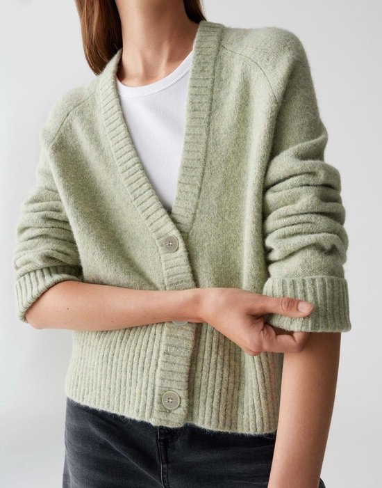 Femme portant un gilet tricoté vert clair à boutons et un haut blanc. Elle remonte sa manche.