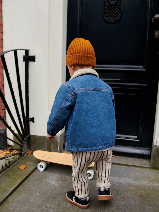 Jongen draagt blauw denim spijkerjack met borgkraag, okergele muts en gestreepte broek.