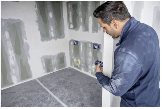 Man in donkerblauwe jas met afstandsmeter in bouwruimte met pleisterwerk.