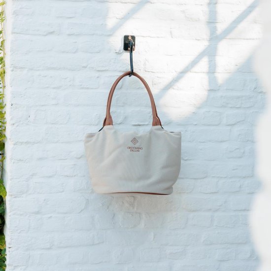 Beige canvas tas met bruine leren hengsels, hangend aan een muur. "GROOMING DELUXE" logo zichtbaar.