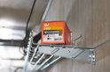 Een rode kartonnen doos met de tekst 'Spit FIX3' bovenop een metalen rail, naast een elektrische kast.