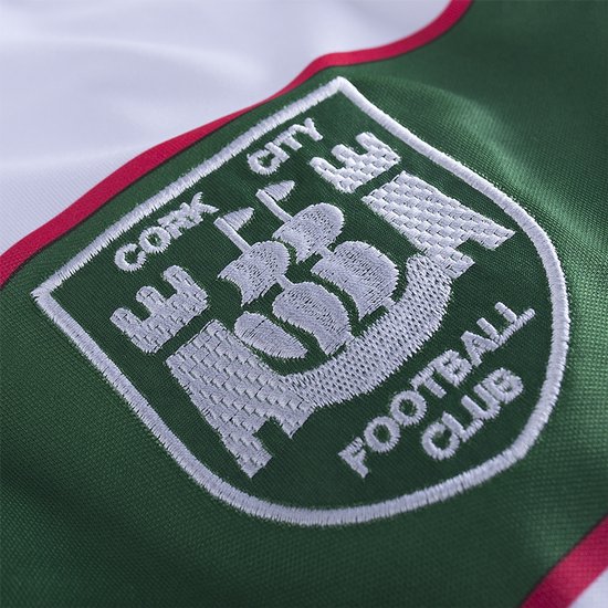 Groen clublogo van Cork City Football Club met schip en gebouwen, geborduurd op een wit shirt.