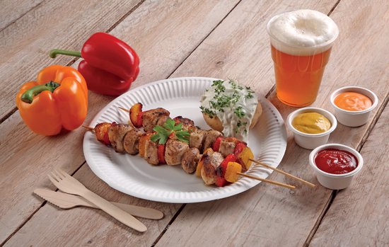 Een grillspies met vlees en groenten op een wit kartonnen bord, naast paprikas, aardappel en bier.