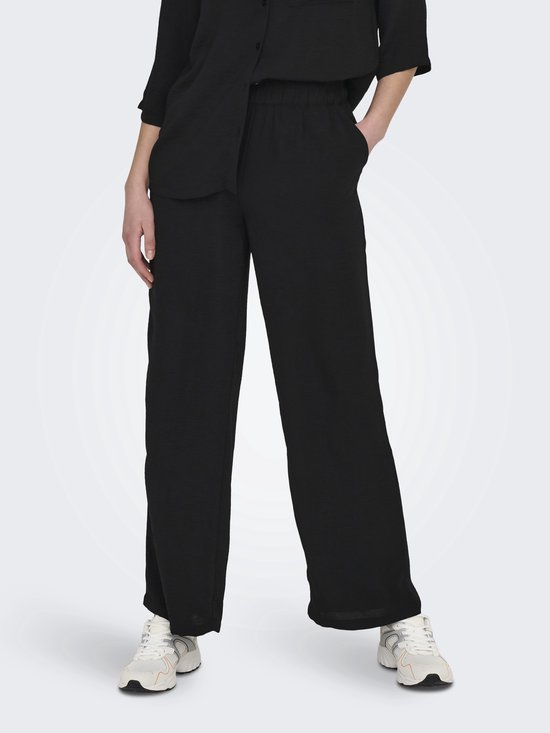 Pantalon noir large avec ceinture élastique et poches latérales, porté par un mannequin avec des baskets blanches.