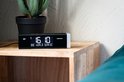 Zwarte Lenco wekkerradio met tijd '16:10', zender 'BBC WORLD SERVICE' op een houten nachtkastje naast een plant.