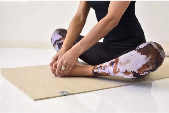 Een vrouw zit in kleermakerszit op een zandkleurige yogamat, met een broek met bruine en paarse abstracte print.