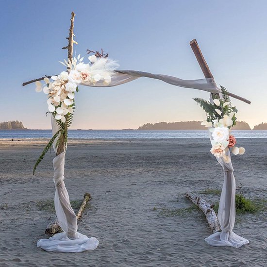Bruiloftboog met zachtroze en witte rozen, eucalyptusbladeren en pampasgras op een strand.