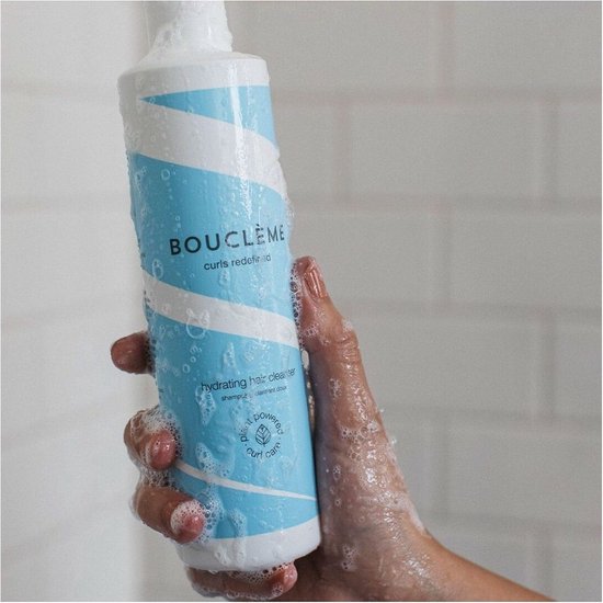 Hand met manicuur houdt een fles hydraterende haarreiniger met blauwe en witte strepen vast, bedekt met waterdruppels en schuim.