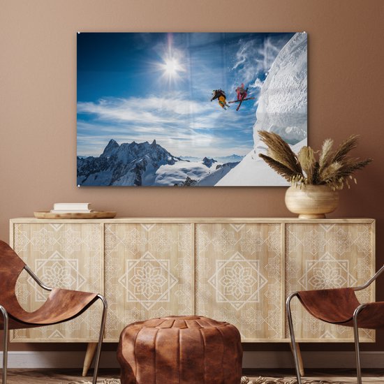 Twee skiërs maken een sprong van een besneeuwde bergtop met een heldere blauwe lucht en zon.