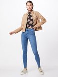Model in een beige jasje, zwarte top met bloemenprint en lichtblauwe skinny jeans. Sneakers.