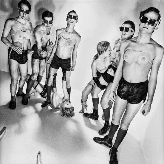 Groep van zes mannen en een vrouw, gekleed in retro-futuristische outfits met vierkante brillen en lichte beha's.