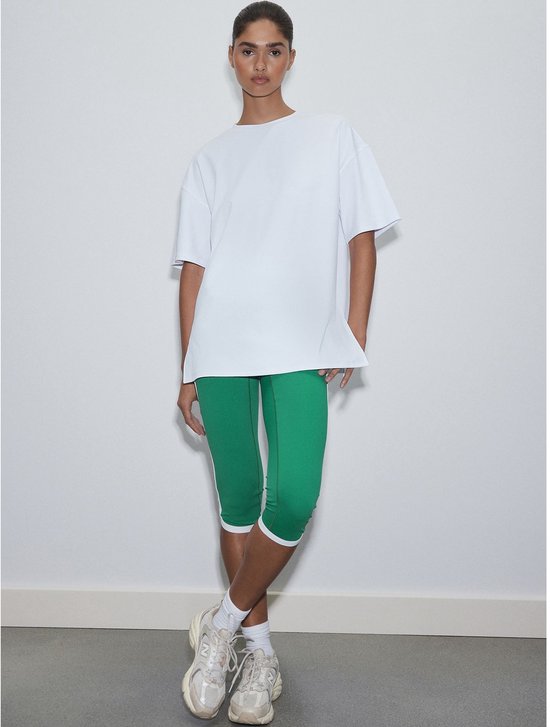 Model draagt een oversized wit T-shirt met driekwart groene sportlegging met witte strepen.