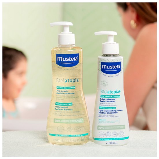 Mustela Stelatopia Cleansing Oil (500ml) naast Stelatopia+ Cream (300ml) met baby op achtergrond.