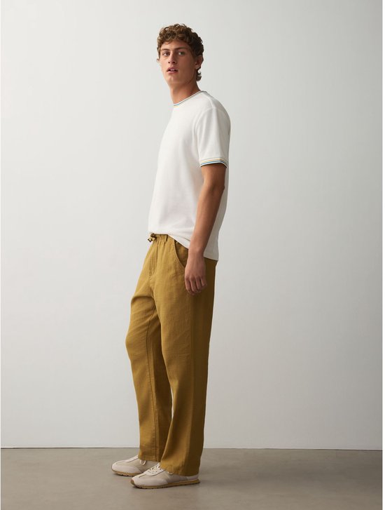 Zijaanzicht van model in okergele broek van linnenmix en witte t-shirt, staand met een ontspannen houding.