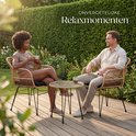 Een man en vrouw genieten van koffie in een tuin op een bruine rotan stoel en glazen tafel, met zonsondergang op de achtergrond.