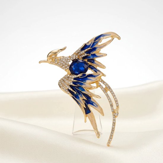 Gouden broche in de vorm van een vogel met blauwe en witte vleugels, een grote blauwe steen en kristallen, op een crèmekleurige zijden stof.