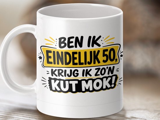 Witte mok met de tekst 'BEN IK EINDELIJK 50, KRIJG IK ZO'N KUT MOK!' op een zachte achtergrond.