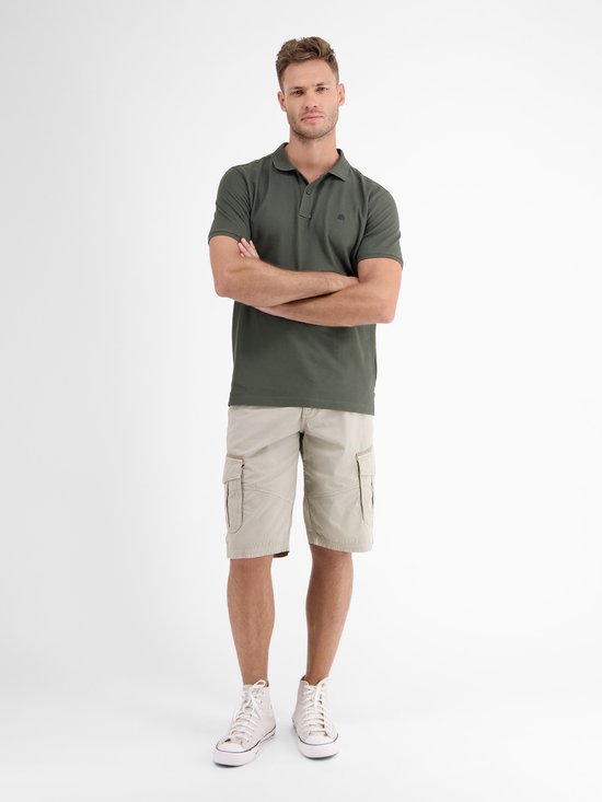 Man met donkergroen poloshirt en beige cargo shorts, staand met armen over elkaar.