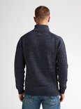 Man met kort bruin haar, draagt een donkerblauwe gebreide trui en spijkerbroek.