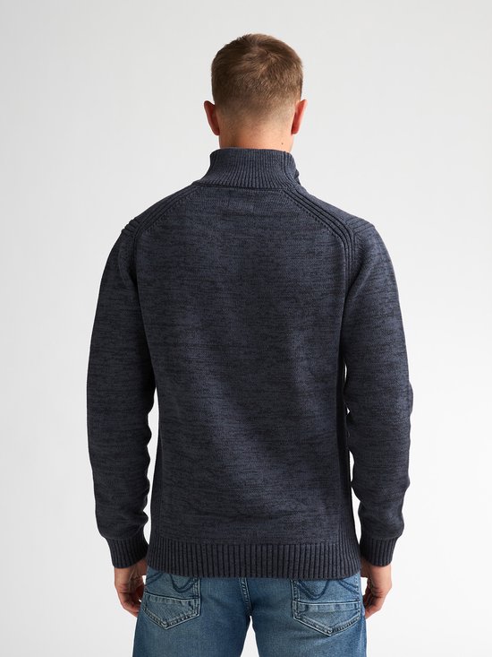 Man met kort bruin haar, draagt een donkerblauwe gebreide trui en spijkerbroek.