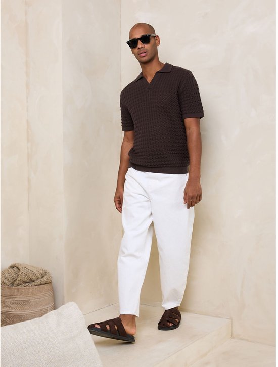 Man met bruin poloshirt, witte broek en sandalen, met zonnebril.