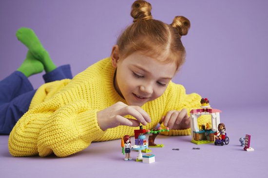 Fille aux couettes rousses et pull jaune jouant avec La Crêperie LEGO Friends sur fond violet.
