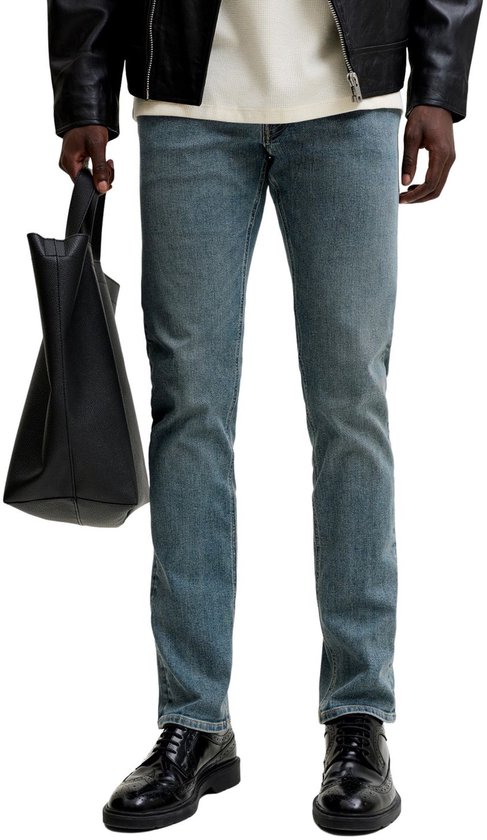 Onderlichaam van een man in blauwe slim fit jeans, met zwarte schoenen en draagt een zwarte tas.