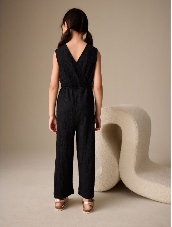 Meisje in zwarte jumpsuit van kreukstof, met V-hals en wijde pijpen. Zichtbaar vanaf de rug.